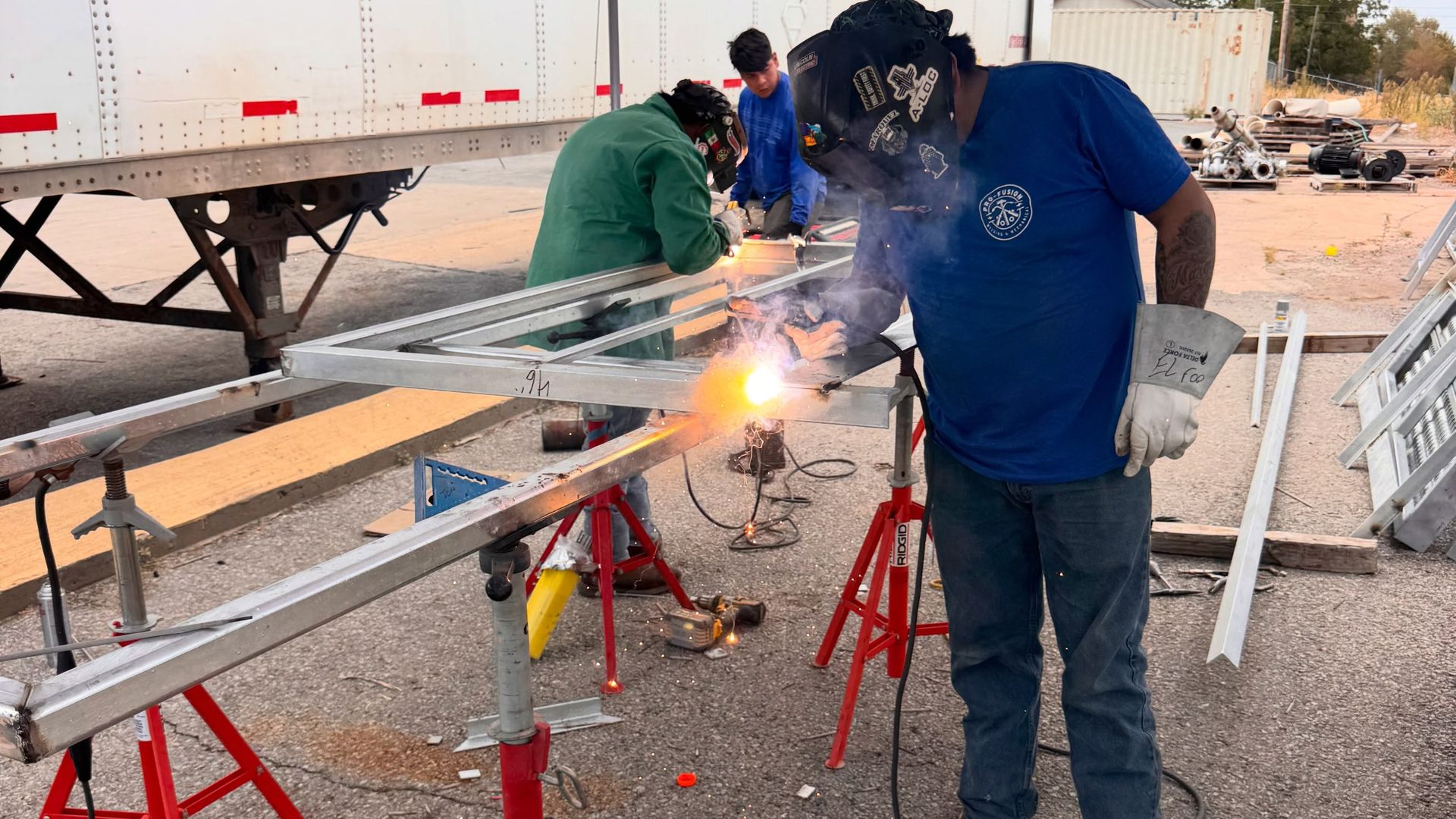 A welder in a blue Pro-fusion shirt welding an aluminum frame on red pipe stands under a canopy, sparks flying off the joint.