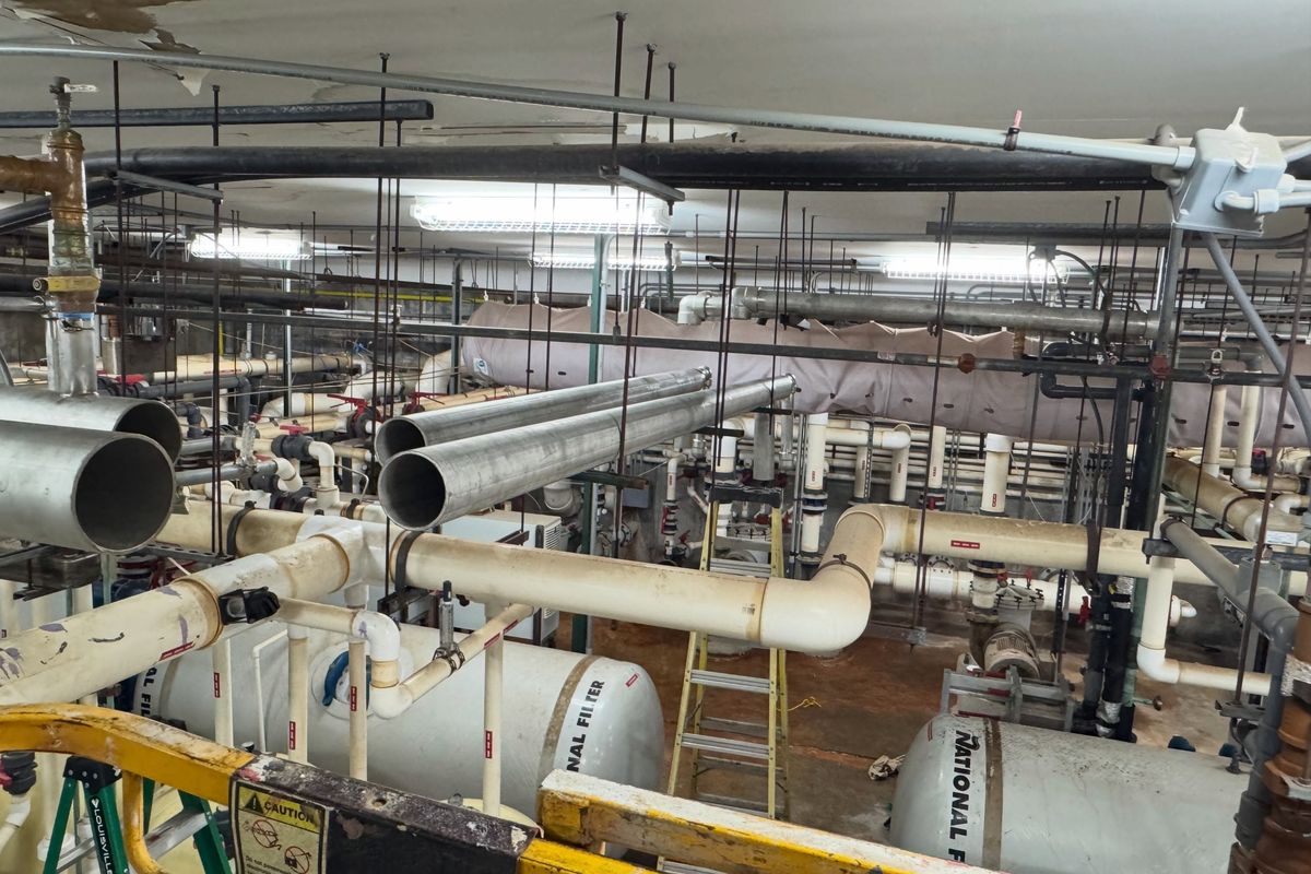 A mechanical room of pipe runs, pressure tanks and a ladder, with new stainless pipe overhead.