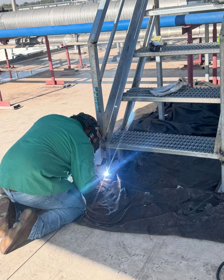 A welder kneeling on a rooftop, welding the base of a galvanized stair, the arc bright blue.