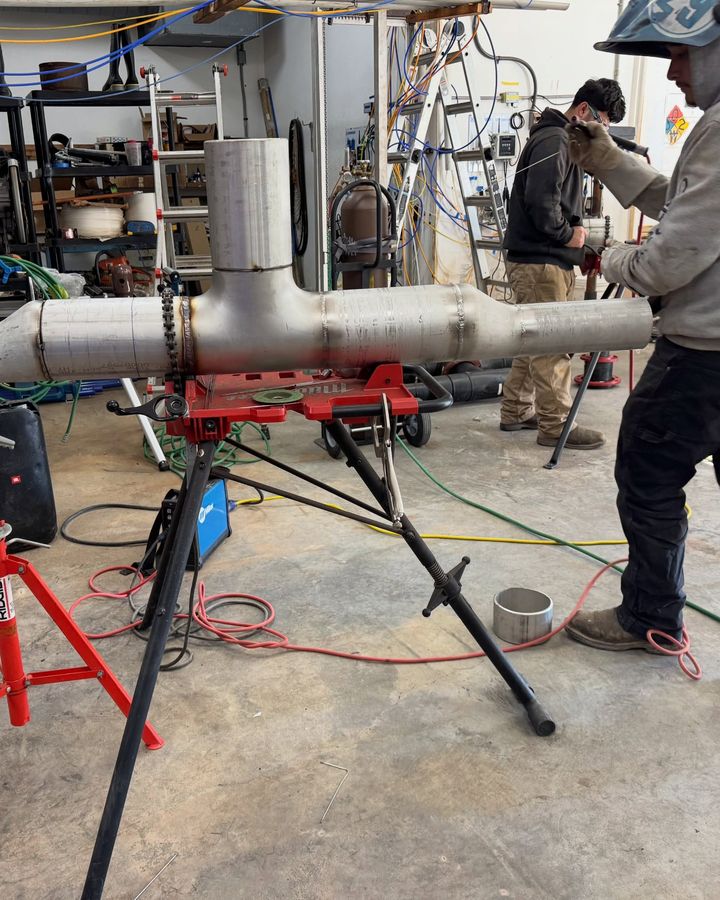 A welder in a shop beside a stainless pipe tee set up on red pipe stands.